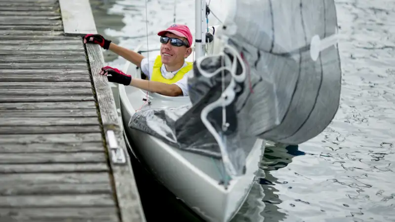 Basti, ein junger Mann mit Sonnenbrille und Basecap, sitzt in einem Segelboot und hält sich am Bootssteg fest.