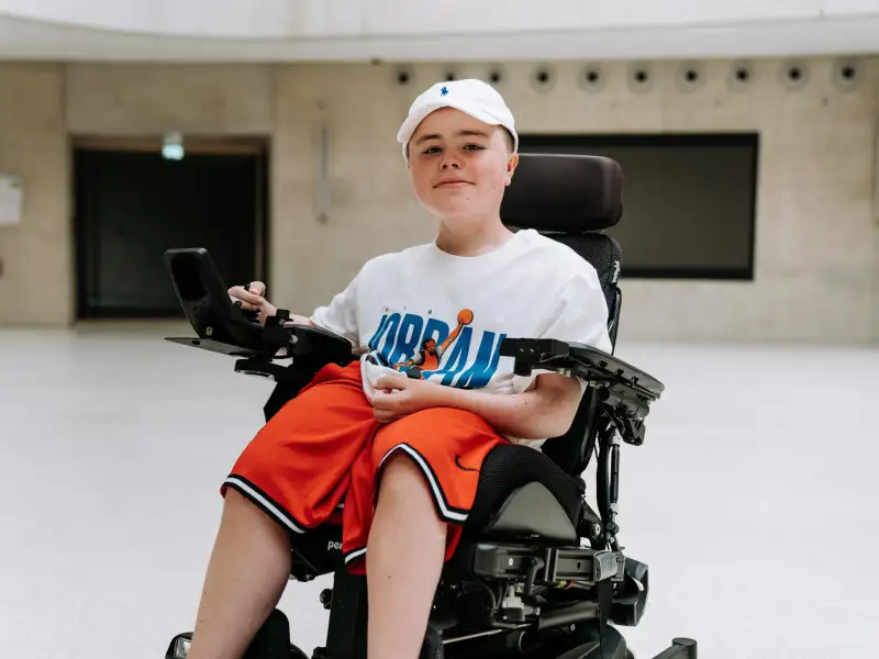 Ein Junge sitzt in einem elektrischen Rollstuhl. Er trägt Basketball-Klamotten und lächelt in die Kamera.