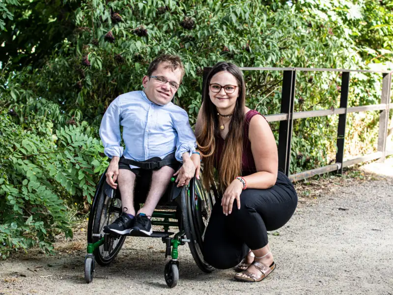 Cristina, eine junge Frau mit langen, dunklen Haaren und Brille hockt neben ihrem Partner, der kurze braune Haare hat, eine Brille trägt und im Rollstuhl sitzt.