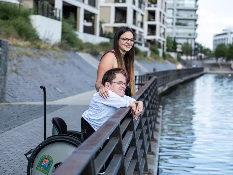 Cristina und Max stehen gemeinsam an einer Uferpromenade und schauen auf den Fluss.