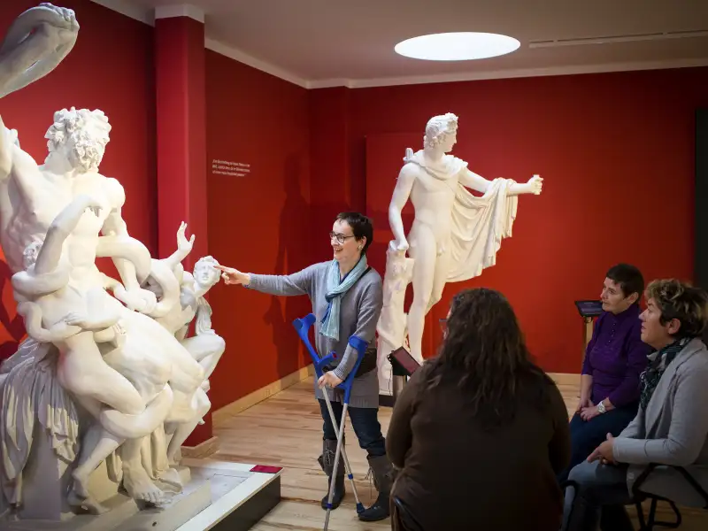 Agnes bei einer Führung im Museum. Sie steht vor einer Statue und zeigt mit dem Finger darauf. Rechts im Bild sitzen Menschen, die ihr zuhören.