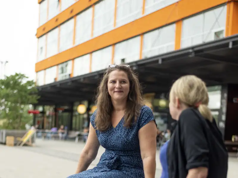 Zwei Frauen sitzen auf einem Mäuerchen und schauen sich an. Eine Frau sieht man von vorne, die andere von hinten. Beide tragen an beiden Beinen Prothesen.