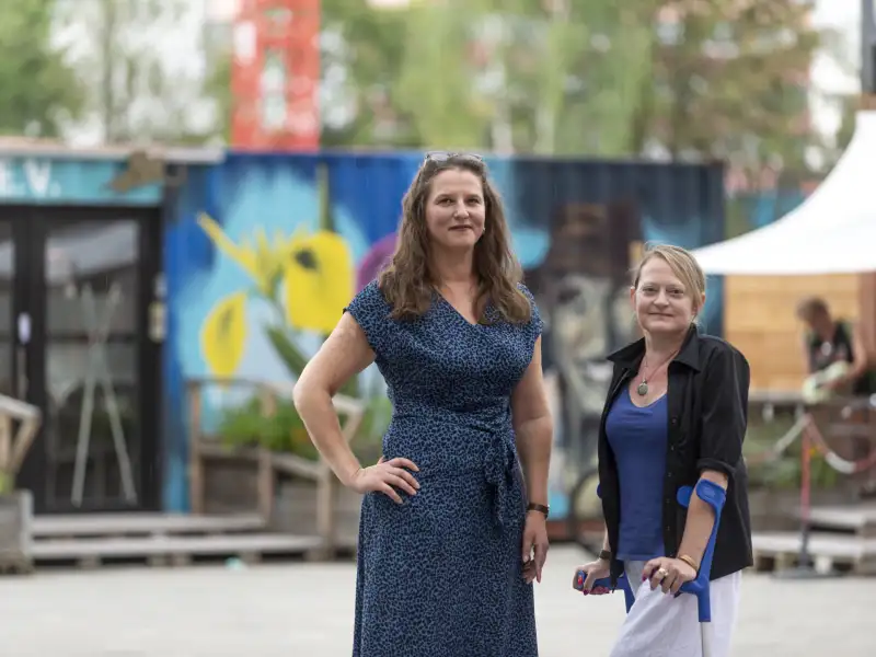 Zwei Frauen stehen nebeneinander auf einem Platz. Eine hat langes, gewelltes Haar, eine Brille auf der Stirn und ist sehr groß. Die andere Frau ist kleiner und hat blondes Haar. Sie nutzt Unterarm-Gestützen. Beide Frauen sieht man nur bis zu den Oberschenkeln.