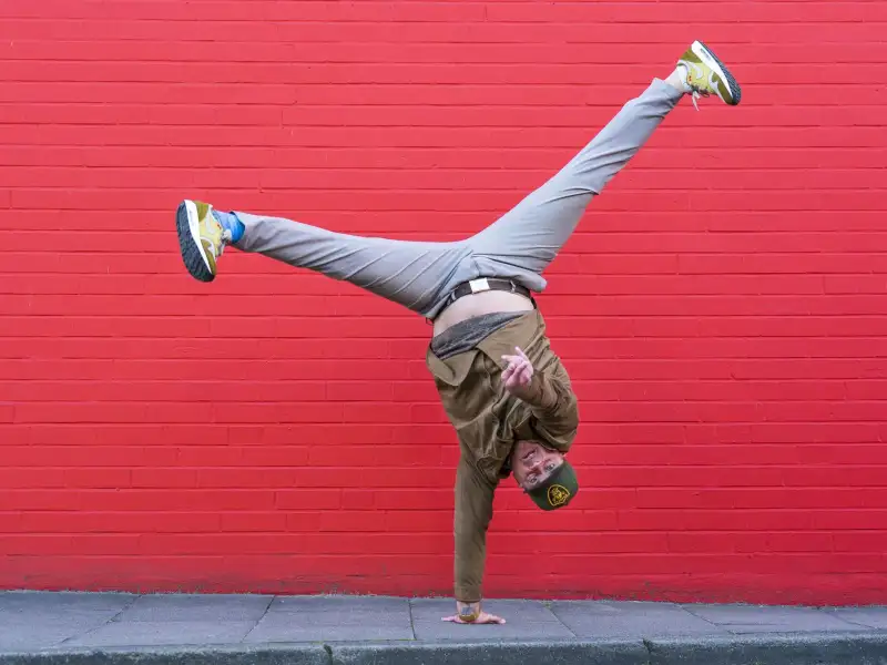 Vor einer rot gestrichenen Wand macht ein Mann einen einhändigen Handstand. Seine Beine sind weit auseinandergespreizt über ihm in der Luft.