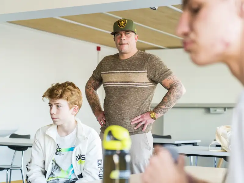 Christian Delles steht in einem Klassenzimmer. Vor ihm sitzen zwei Jugendliche.