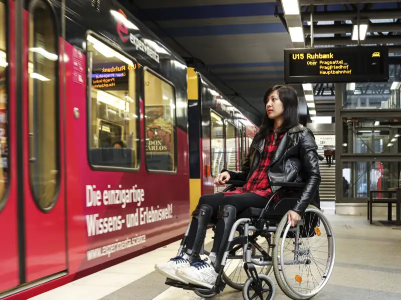 Simone, eine junge Frau mit langen dunklen Haaren im Rollstuhl, steht an einer U-Bahn-Haltestelle. Die Bahn ist gerade eingefahren.