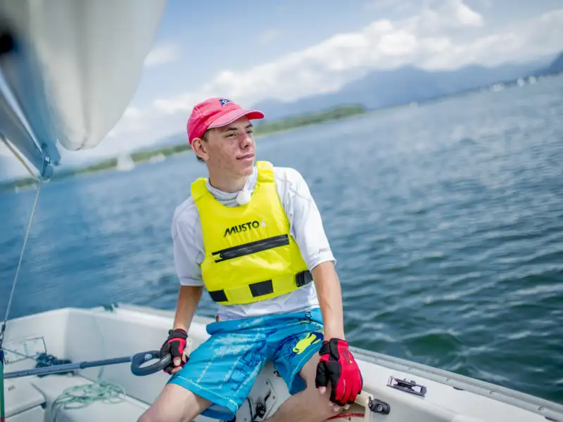 Basti, ein junger Mann mit kurzen Haaren sitzt in einem Segelboot. Er trägt ein rotes Basecap, ein weißes Shirt, blaue Shorts und eine Schwimmweste. Im Hintergrund sieht man einen See.