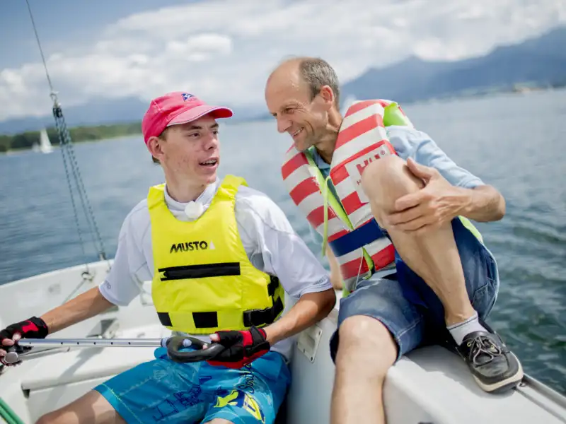 Basti, ein junger Mann mit Basecap und ein Mann sitzen nebeneinander auf einem Segelboot. Beide tragen Schwimmwesten. Im Hintergrund sieht man einen See.