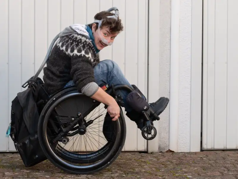 Eine junge Frau balanciert im Rollstuhl. Sie trägt ein Beatmungsgerät.