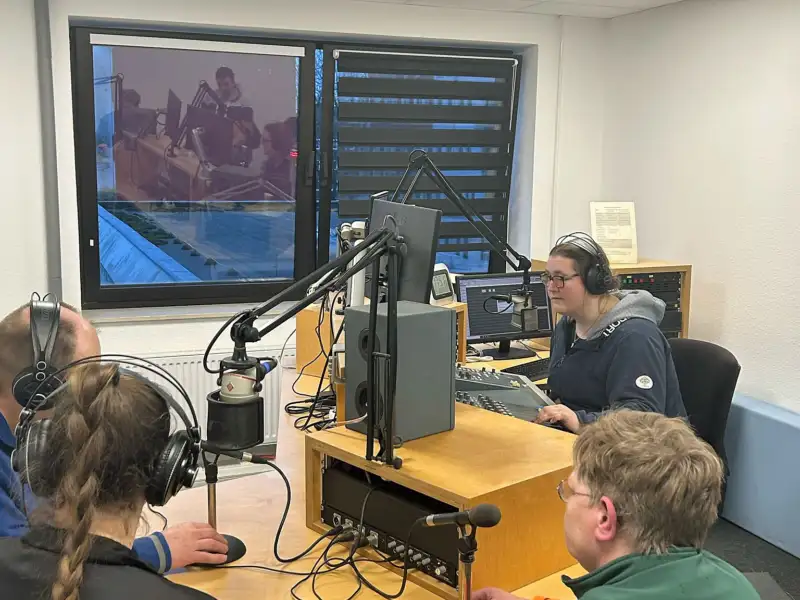 Vier Personen in einem Aufnahmestudio. Vor ihnen stehen Mikrofone. Einige tragen Kopfhörer.