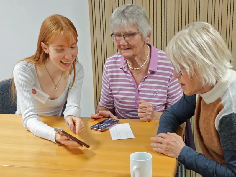 Eine junge Frau mit langem rötlichen Haar zeigt zwei Seniorinnen etwas auf einem Smartphone.