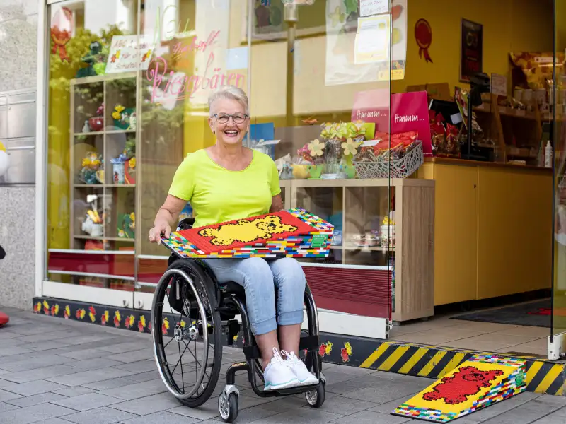 Eine Frau mit kurzen grauen Haaren und sommerlicher Kleidung sitzt in ihrem Rollstuhl vor einem Süßwarenladen und hält eine ihrer selbstgebauten Legorampen auf dem Schoß. Auf der Rampe sind bunte Gummibärchen abgebildet.