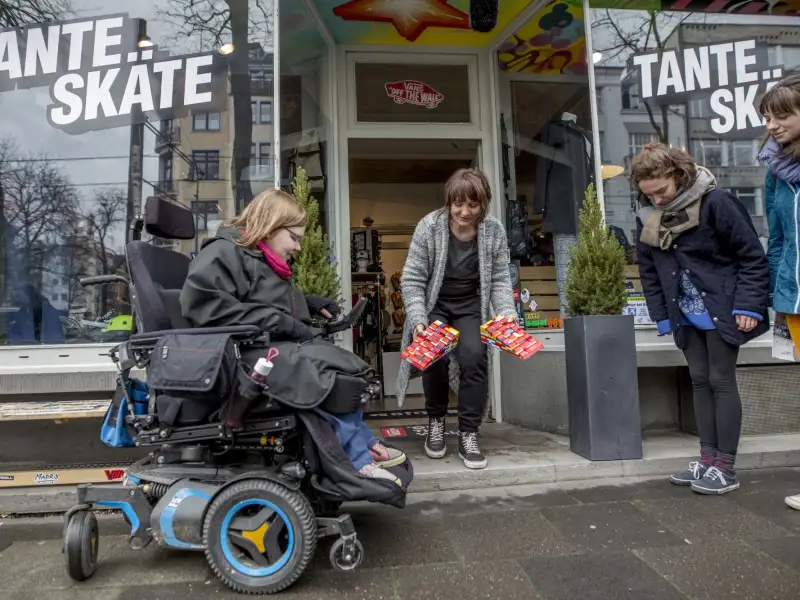 Vier Menschen stehen vor dem Eingang eines Ladens. Eine Person sitzt im Rollstuhl und schaut auf die Eingangsstufe. Eine Frau hat zwei Rampen aus Legosteinen in den Händen. Sie steht im Eingang und beugt sich zur Stufe herunter.
