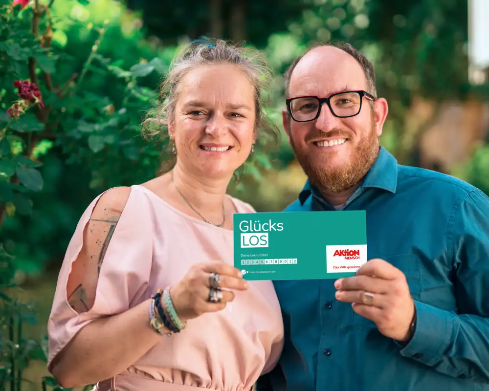 Gewinner-Ehepaar Adriana und Benjamin stehen im Garten und lachen in die Kamera. Gemeinsam halten sie ein Glücks-Los in ihren Händen.