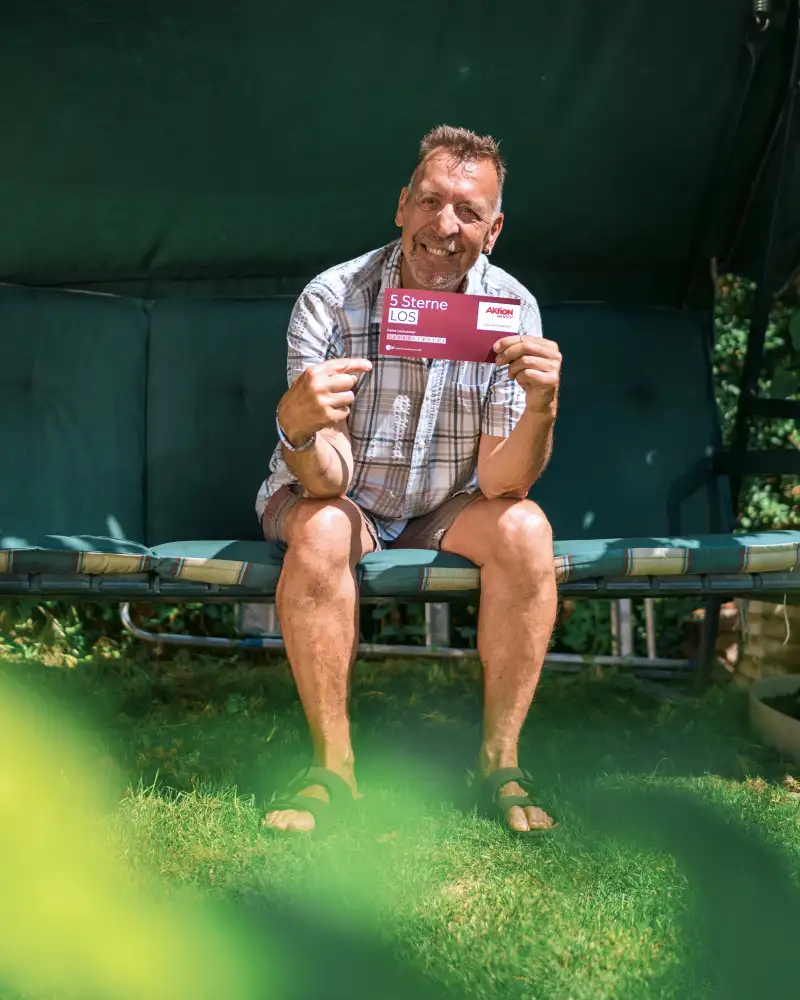 Aktion Mensch-Gewinner Ralf sitzt in seiner Hollywood-Schaukel. In der Hand hält er ein Aktion Mensch-5-Sterne-Los.