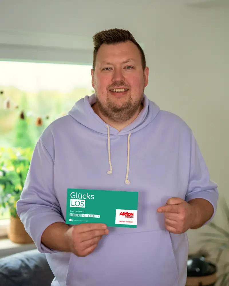 Der Aktion Mensch-Gewinner Finn steht in seinem Wohnzimmer. In der rechten Hand hält er ein Aktion Mensch-Glücks-Los. Mit dem linken Zeigefinger zeigt er auf das Los.