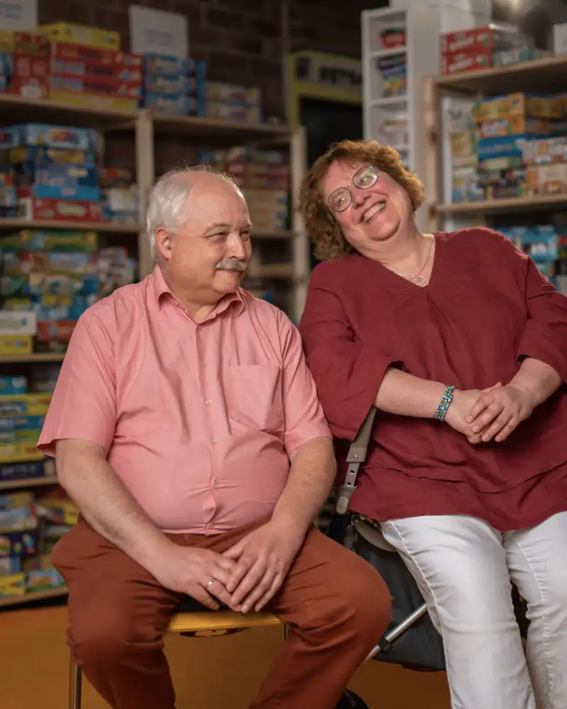 Die Aktion Mensch-Gewinner*innen Claudia und Michael. Im Hintergrund sieht man Regale. In diesen Regalen sind zahlreiche Gesellschaftsspiele.