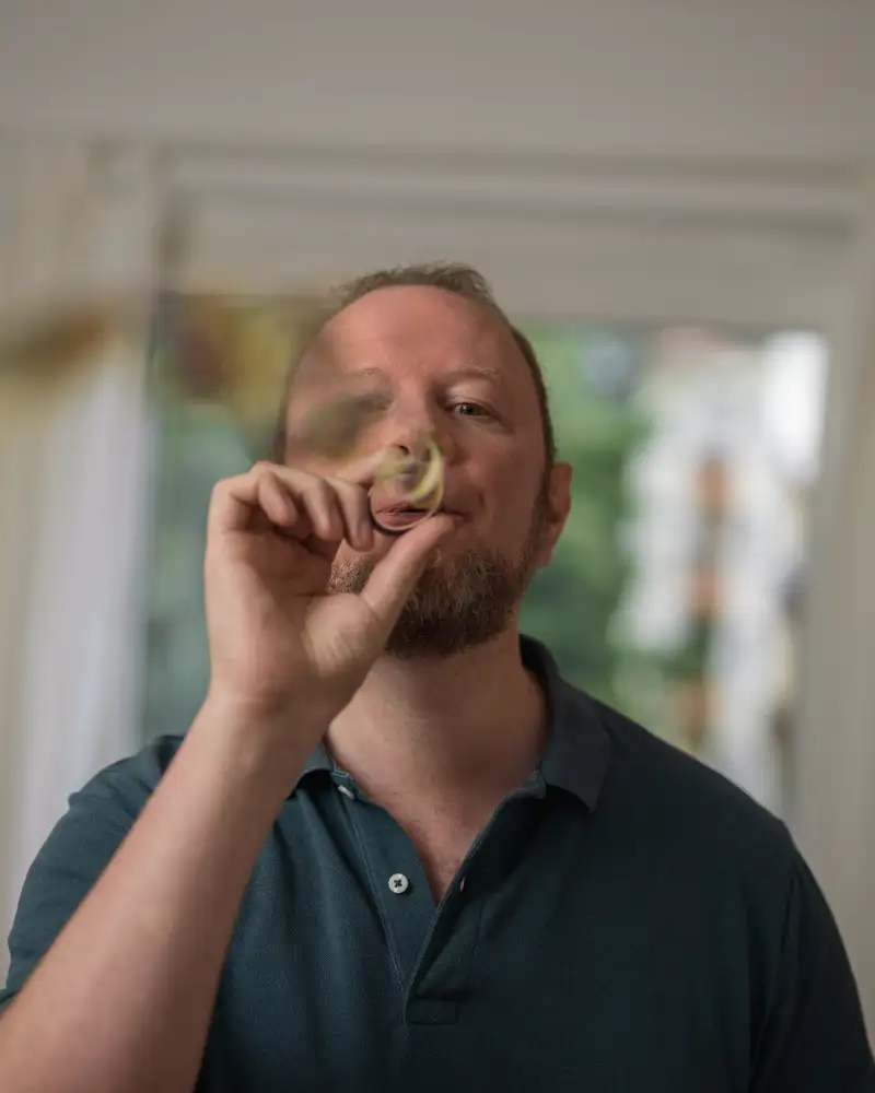 Aktion Mensch-Gewinner Bastian steht in seinem Wohnzimmer und pustet in eine Luftschlange.