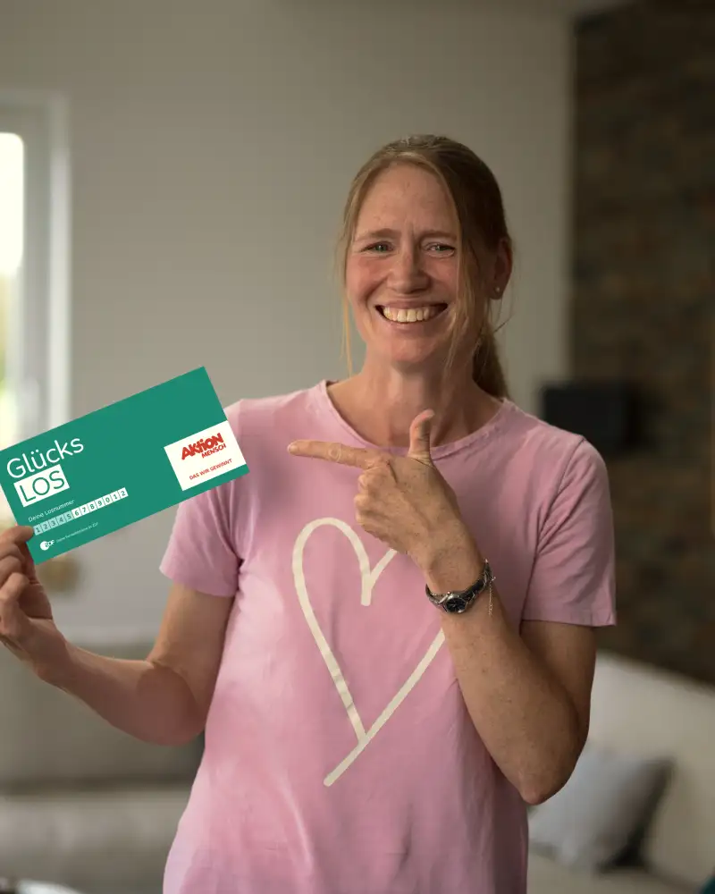 Aktion Mensch-Gewinnerin Stefanie steht in ihrem Wohnzimmer. In der rechten Hand hält sie ein Aktion Mensch Glücks-Los. Mit der linken Hand zeigt sie auf dieses Los.