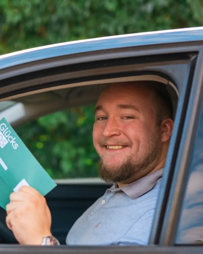 Gewinner David sitzt in seinem neuen Auto. Das Fenster an der Fahrerseite ist offen und er hält lächelnd zwei Glücks-Lose in die Kamera.