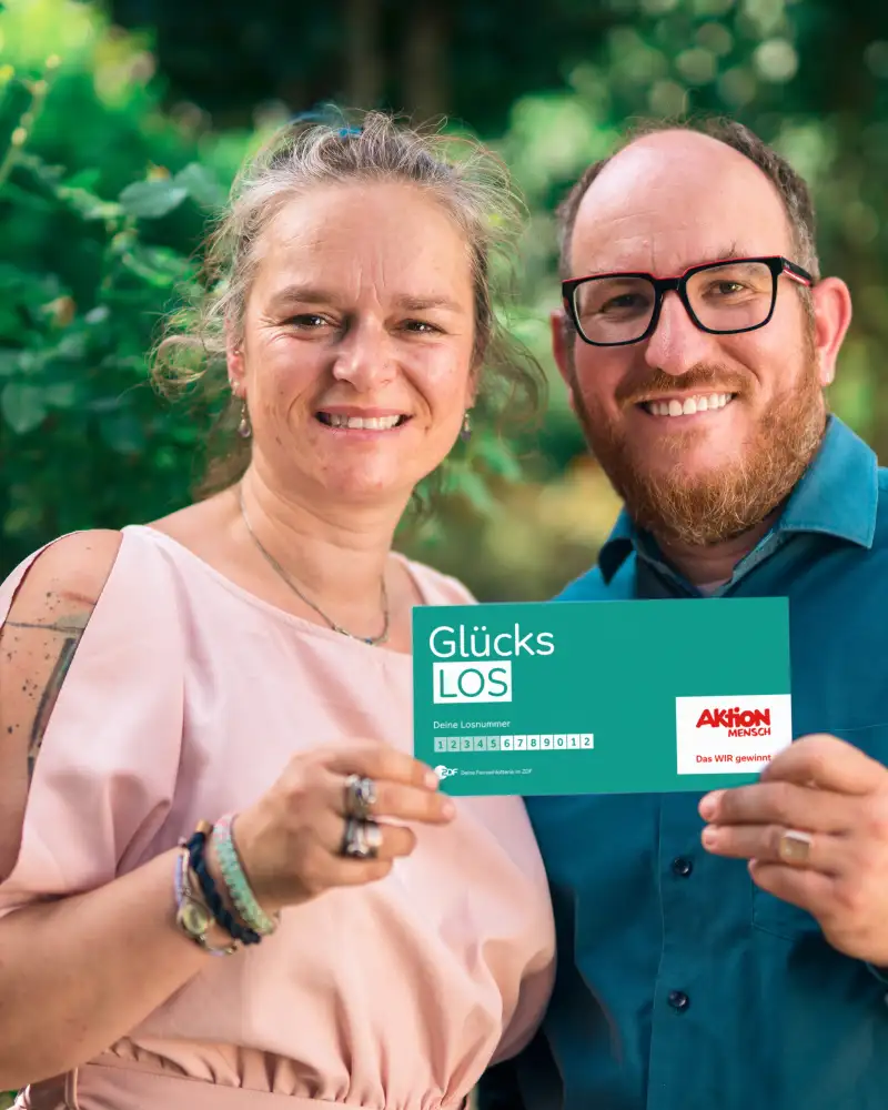 Gewinner-Ehepaar Adriana und Benjamin stehen im Garten und lachen in die Kamera. Gemeinsam halten sie ein Glücks-Los in ihren Händen.