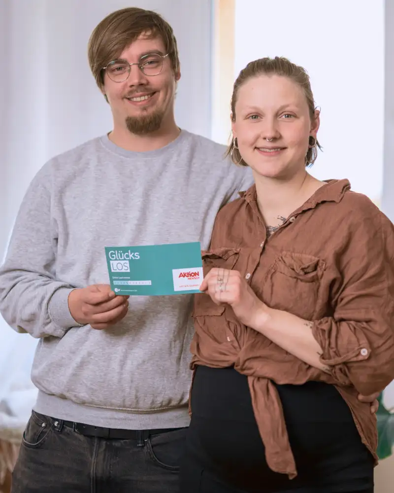 Aktion Mensch-Gewinner Sandro umarmt seine Freundin Fine. Sie stehen in ihrem Wohnzimmer und halten gemeinsam ein Aktion Mensch-Los fest.