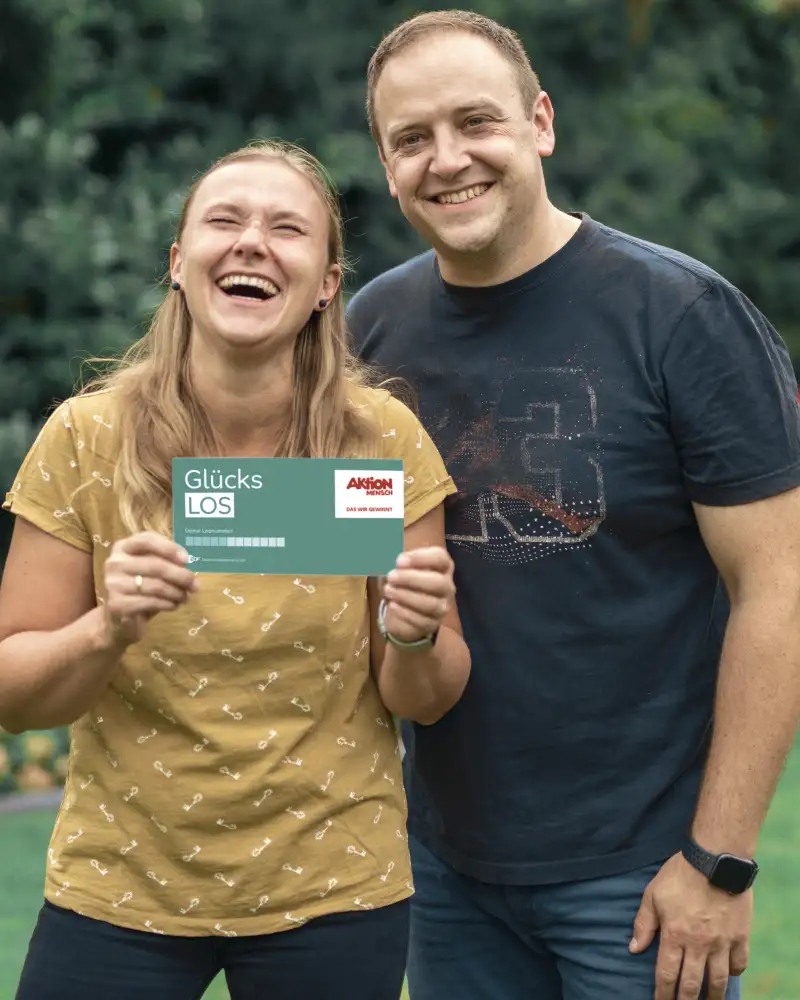 Aktion Mensch-Gewinnerin Bozena mit ihrem Mann im Garten. Sie lachen beide. Bozena hält ein Aktion Mensch-Los in der Hand.