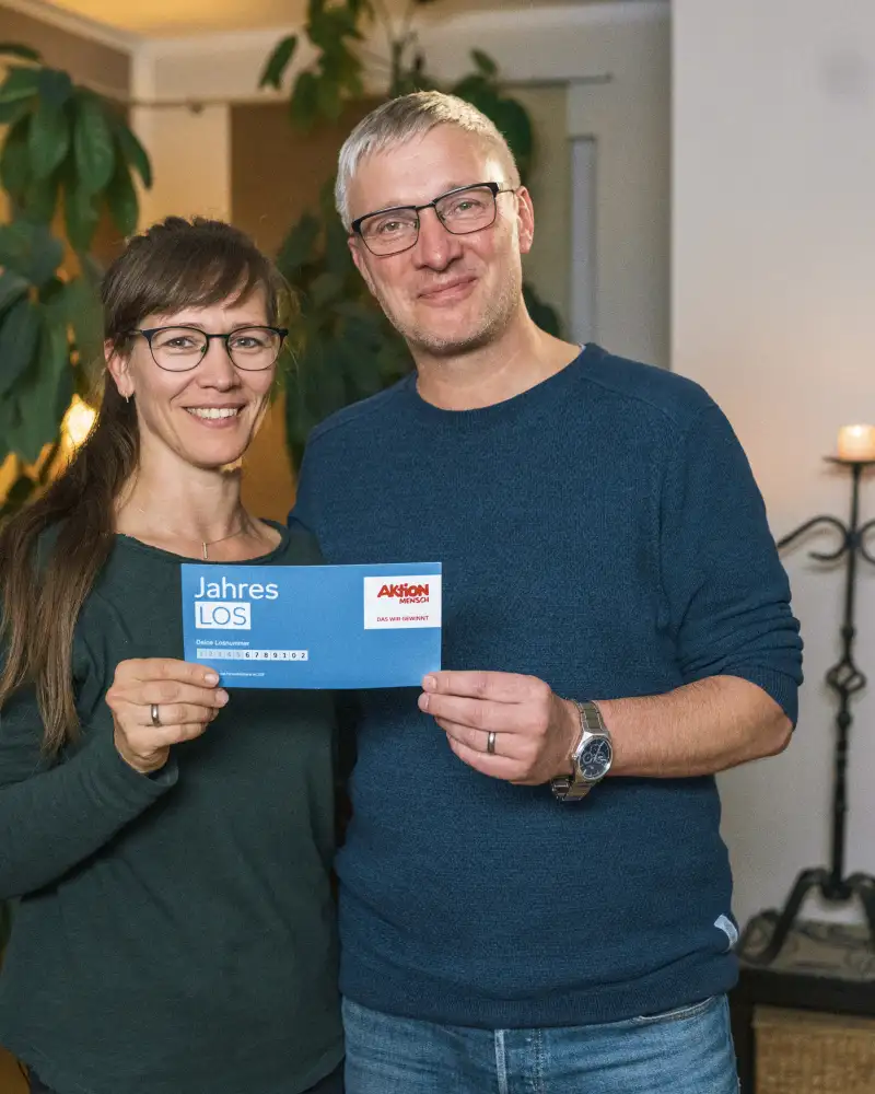 Aktion Mensch-Gewinner Jörg mit seiner Frau Adriane in ihrem Wohnzimmer. Gemeinsam halten sie ein Aktion Mensch-Los.