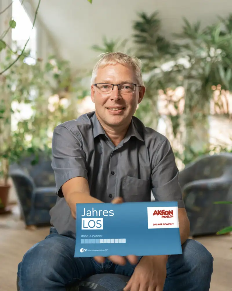 Aktion Mensch-Gewinner Tobias sitzt in seinem Wohnzimmer. Er hält ein Aktion Mensch-Los in die Kamera.
