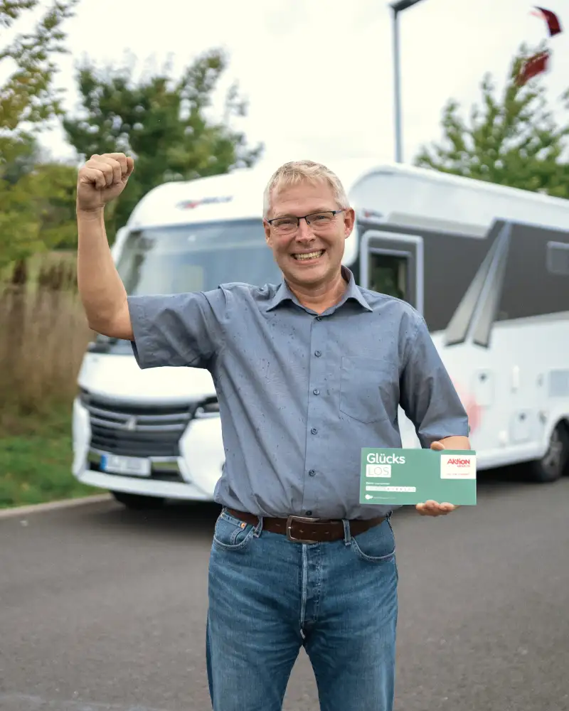 Aktion Mensch-Gewinner Tobias steht jubelnd vor einem Wohnmobil. In einer Hand hält er ein Aktion Mensch-Los.