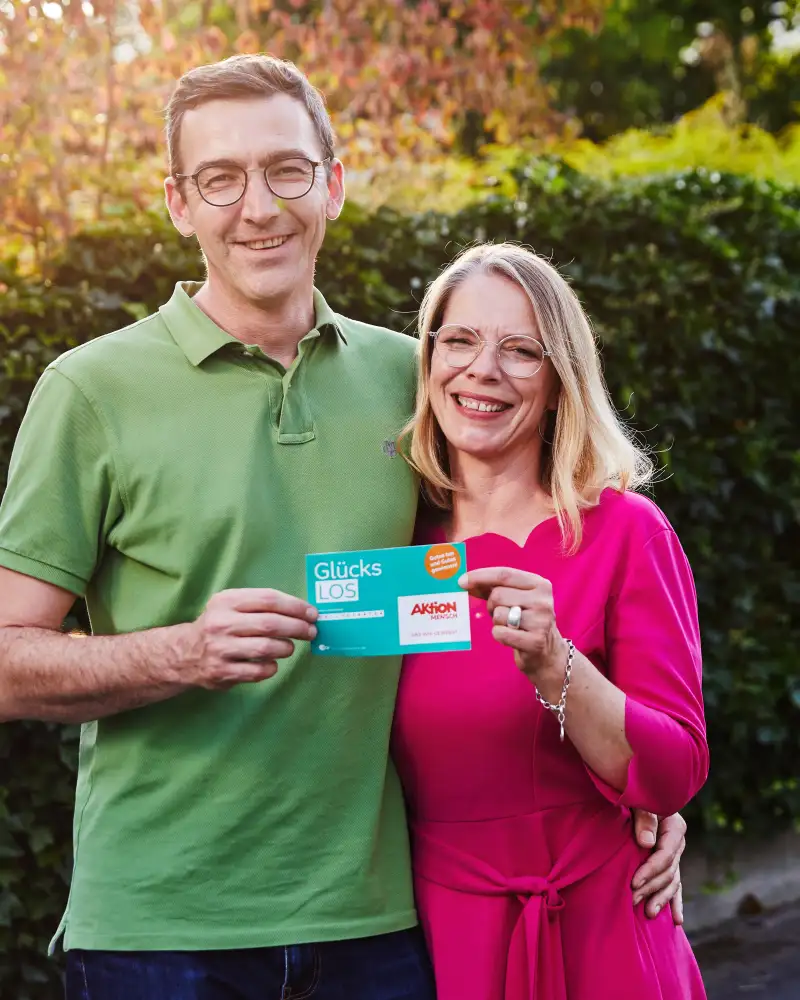 Aktion Mensch-Gewinner Stefan steht mit seiner Frau im Garten. Gemeinsam halten sie ein Aktion Mensch-Los fest.