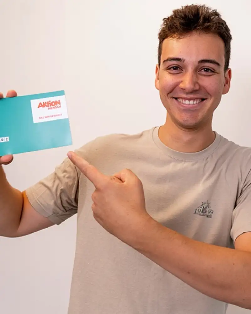 Aktion Mensch-Gewinner Benjamin steht vor einer hellen Wand. In der einen Hand hält er ein Aktion Mensch-Los mit der anderen Hand zeigt er auf dieses Los.