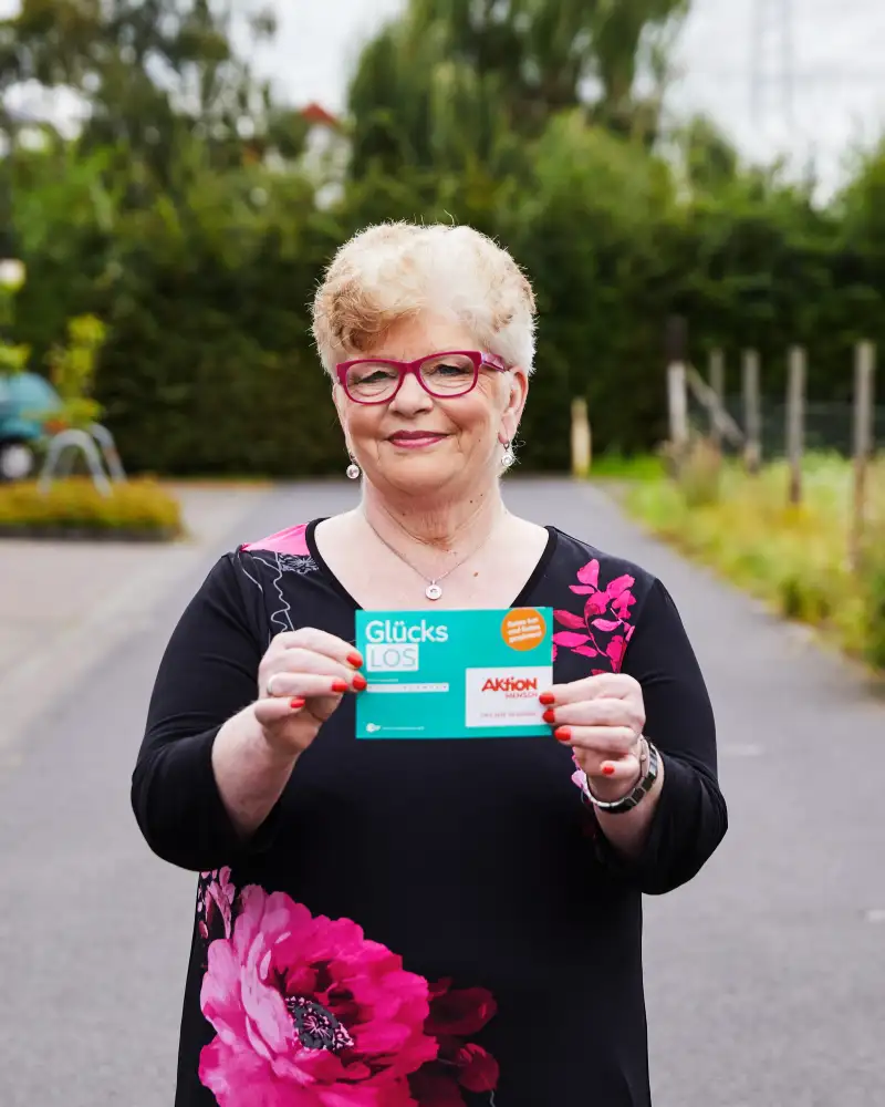 Aktion Mensch-Gewinner Gaby steht auf einer Straße. Sie hält ein Aktion Mensch-Los in der Hand