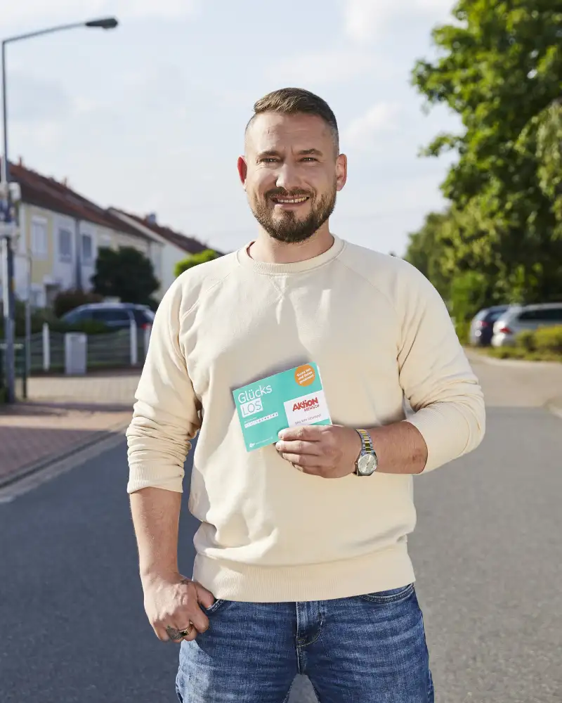 Aktion Mensch-Gewinner Anton steht auf einer Straße und hält ein Aktion Mensch-Los in der Hand.