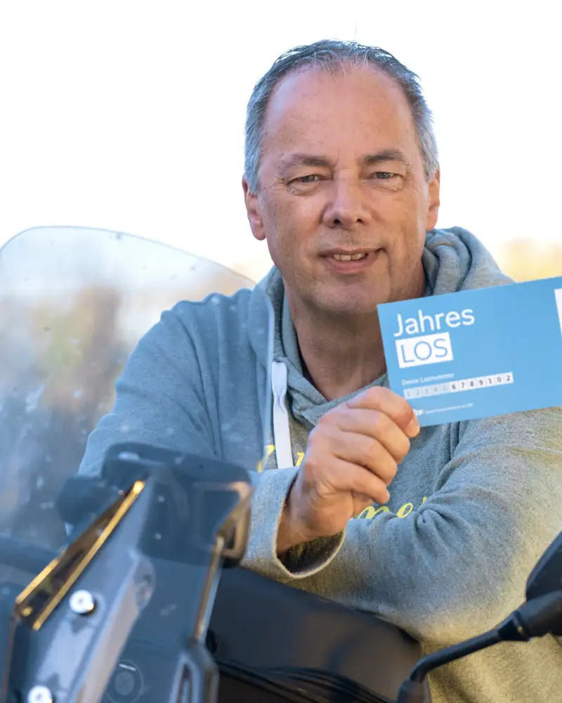 Aktion Mensch-Gewinner Jürgen sitzt auf einem Motorrad und hält ein Aktion Mensch-Los in der Hand.