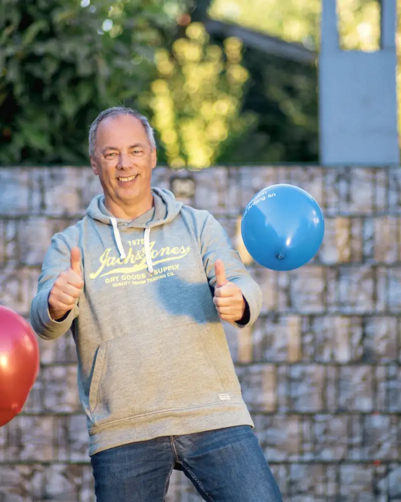 Aktion Mensch-Gewinner Jürgen steht in seinem Garten. Er reckt beide Daumen hoch und es fliegen Luftballons.