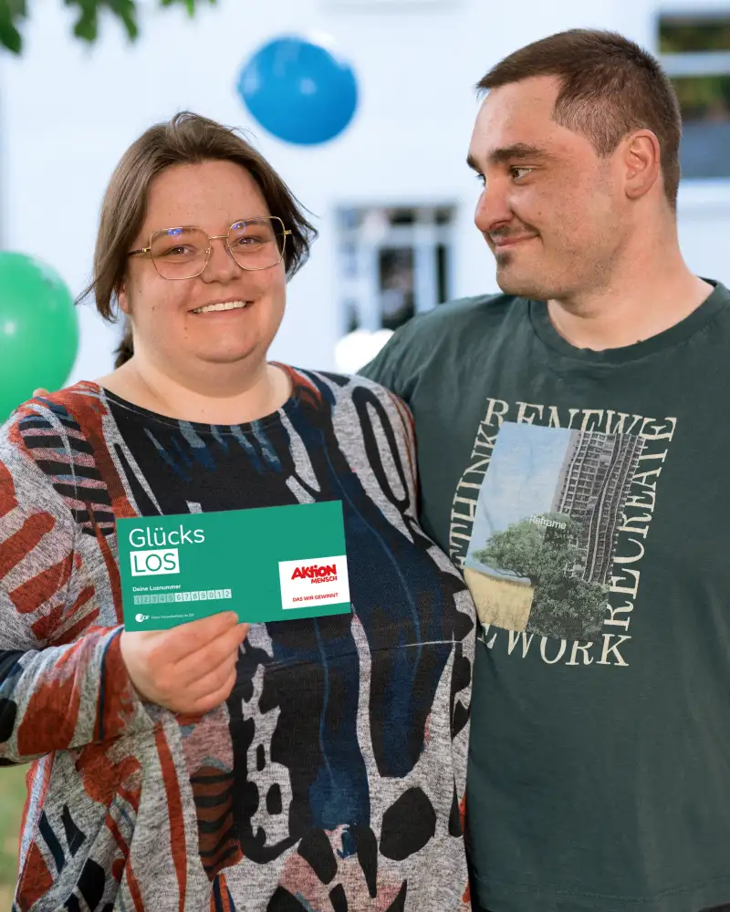 Aktion Mensch-Gewinner Felix und seine Frau Luisa vor einem Haus. Luisa hält ein Aktion Mensch-Los in der Hand. Im Hintergrund schweben Luftballons.