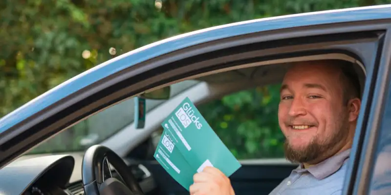 Gewinner David sitzt in seinem neuen Auto. Das Fenster an der Fahrerseite ist offen und er hält lächelnd zwei Glücks-Lose in die Kamera.