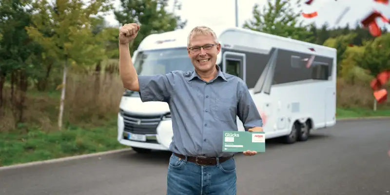 Aktion Mensch-Gewinner Tobias steht jubelnd vor einem Wohnmobil. In einer Hand hält er ein Aktion Mensch-Los.