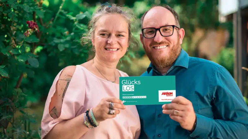 Gewinner-Ehepaar Adriana und Benjamin stehen im Garten und lachen in die Kamera. Gemeinsam halten sie ein Glücks-Los in ihren Händen.