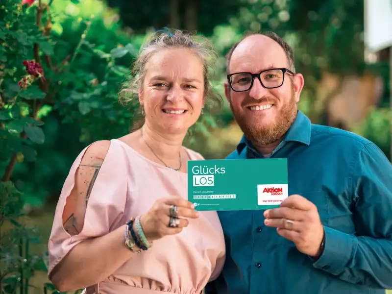 Gewinner-Ehepaar Adriana und Benjamin stehen im Garten und lachen in die Kamera. Gemeinsam halten sie ein Glücks-Los in ihren Händen.