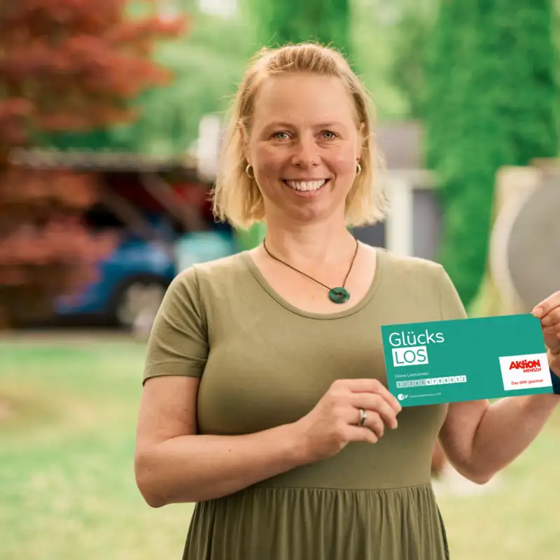 Aktion Mensch-Gewinnerin Claudia, eine Frau mit kinnlangen, blonden Haaren, steht in ihrem Garten und lächelt. In ihren Händen hält sie ein Aktion Mensch-Los.
