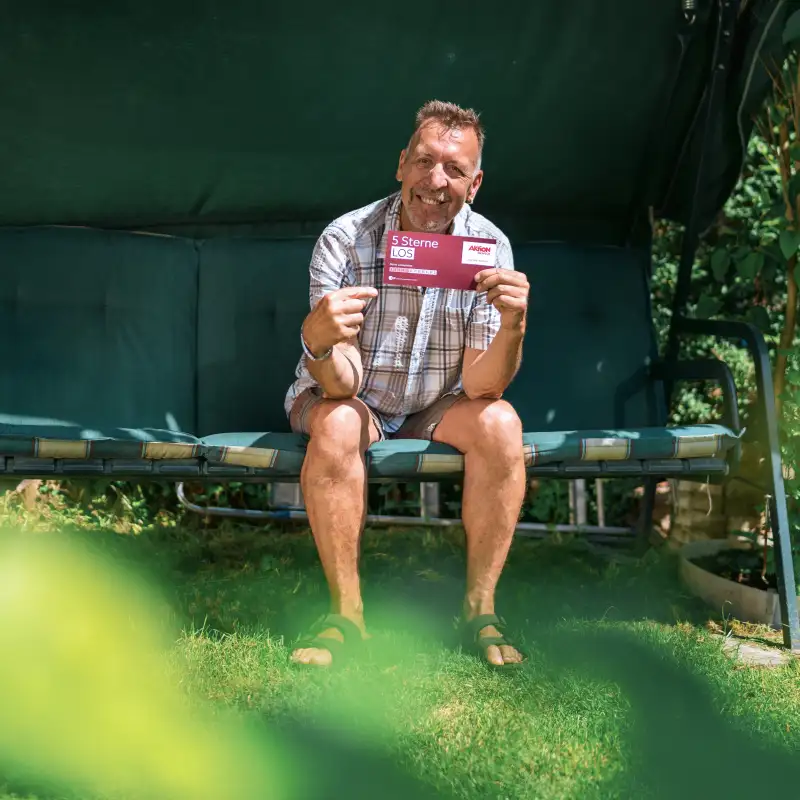 Aktion Mensch-Gewinner Ralf sitzt in seiner Hollywood-Schaukel. In der Hand hält er ein Aktion Mensch-5-Sterne-Los.