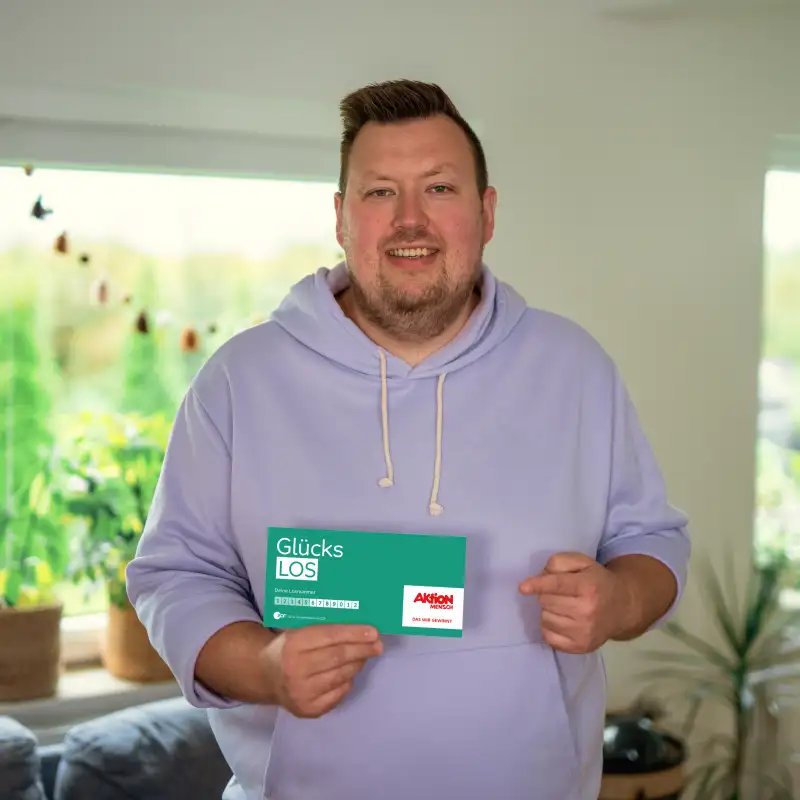 Der Aktion Mensch-Gewinner Finn steht in seinem Wohnzimmer. In der rechten Hand hält er ein Aktion Mensch-Glücks-Los. Mit dem linken Zeigefinger zeigt er auf das Los.