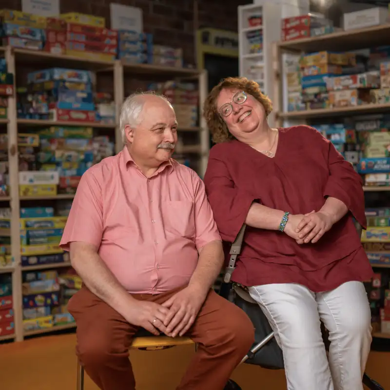 Die Aktion Mensch-Gewinner*innen Claudia und Michael. Im Hintergrund sieht man Regale. In diesen Regalen sind zahlreiche Gesellschaftsspiele.