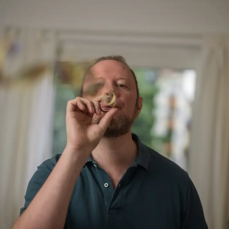 Aktion Mensch-Gewinner Bastian steht in seinem Wohnzimmer und pustet in eine Luftschlange.