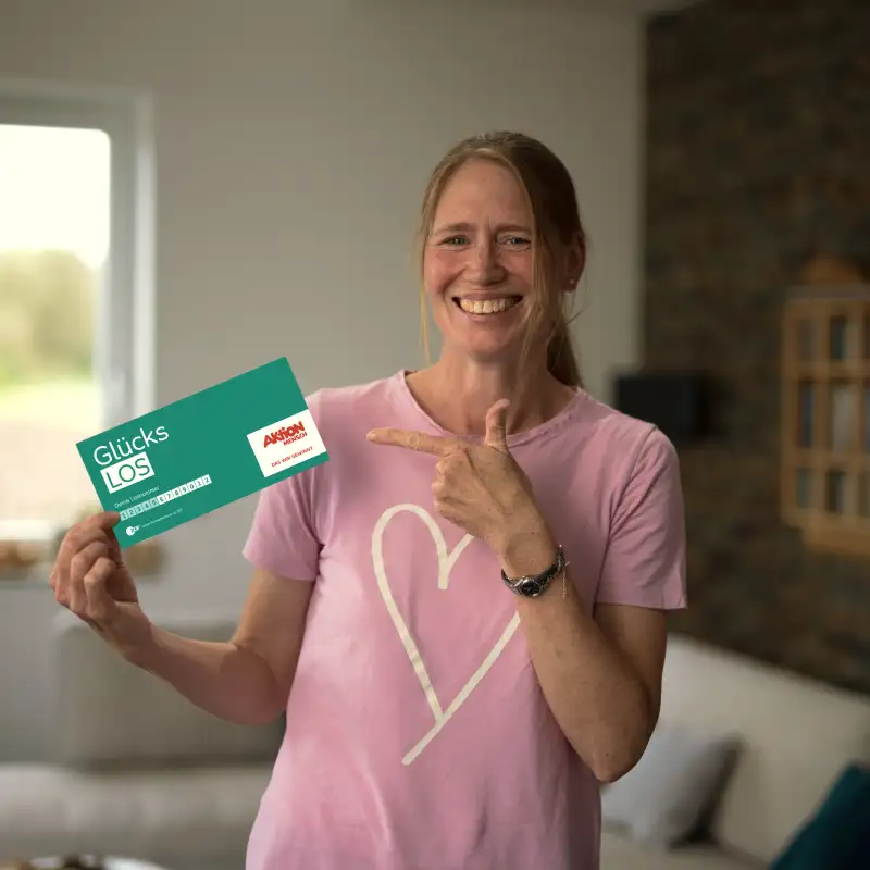 Aktion Mensch-Gewinnerin Stefanie steht in ihrem Wohnzimmer. In der rechten Hand hält sie ein Aktion Mensch Glücks-Los. Mit der linken Hand zeigt sie auf dieses Los.