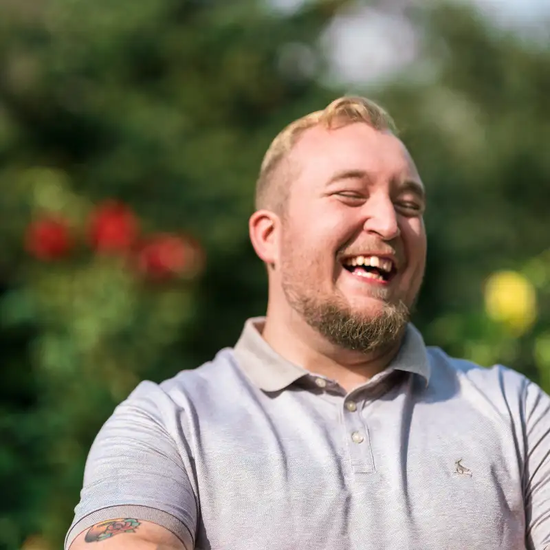 Aktion Mensch-Gewinner David, ein junger Mann mit kurzen, blonden Haaren und Bart, steht in einem Garten und lacht herzhaft.