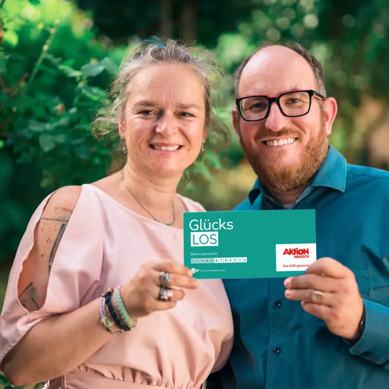 Gewinner-Ehepaar Adriana und Benjamin stehen im Garten und lachen in die Kamera. Gemeinsam halten sie ein Glücks-Los in ihren Händen.