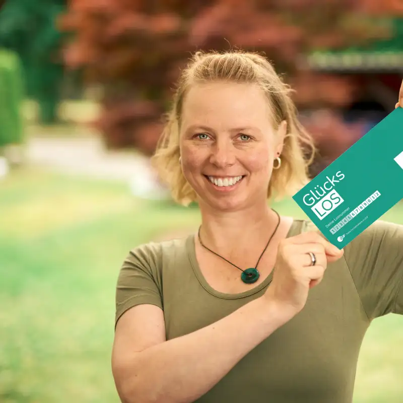 Aktion Mensch-Gewinnerin Claudia, eine Frau mit kinnlangen, blonden Haaren, steht in ihrem Garten und lächelt. In ihren Händen hält sie ein Aktion Mensch-Los in die Höhe.